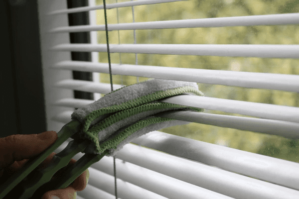 A close-up of a person cleaning white Venetian blinds using a specialized microfiber brush for effective dust removal.