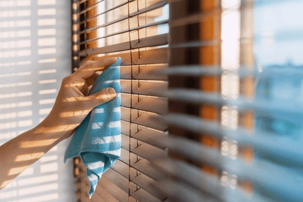 A hand using a blue cloth to clean stylish wooden blinds in bright sunlight, showcasing the best and cheap wooden blinds 2025 in Dubai.