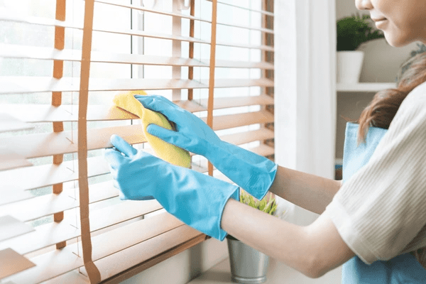 A woman wearing blue gloves and an apron wipes wooden blinds with a yellow cloth, showcasing an easy DIY blind cleaning method in Dubai 2025.