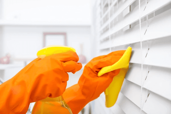 A person wearing orange gloves cleaning white roman blinds with a yellow cloth and spray bottle, ensuring spotless blinds in Dubai 2025.