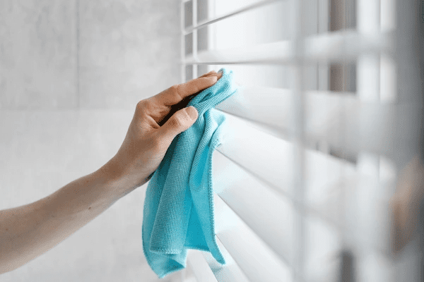 A close-up of a hand wiping white blinds with a blue microfiber cloth, demonstrating a simple DIY blind cleaning method in Dubai 2025.