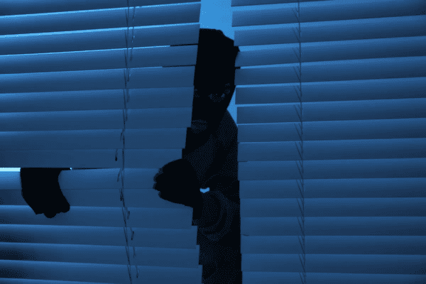 A masked figure peeking through broken blinds in a dark room, symbolizing scams and fraud in the Dubai window blinds market.