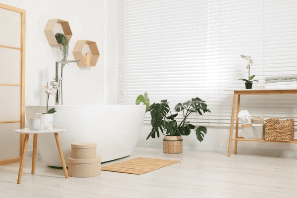A serene and modern bathroom with a white freestanding bathtub, wooden accents, lush green plants, and stylish white window blinds, offering both privacy and elegance.