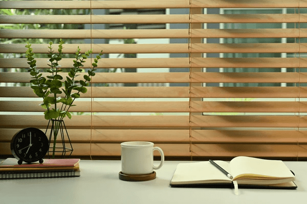 A modern workspace with elegant wooden bay window blinds, a notebook, coffee cup, and houseplant on a desk.