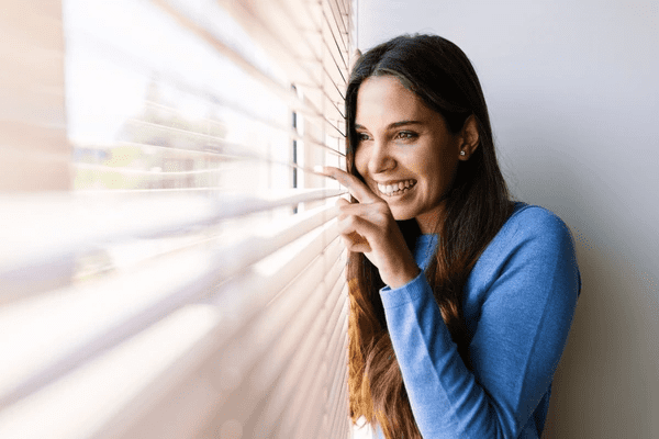 A smiling woman in a blue sweater peeking through elegant bay window blinds, enjoying the natural light and cozy ambiance.