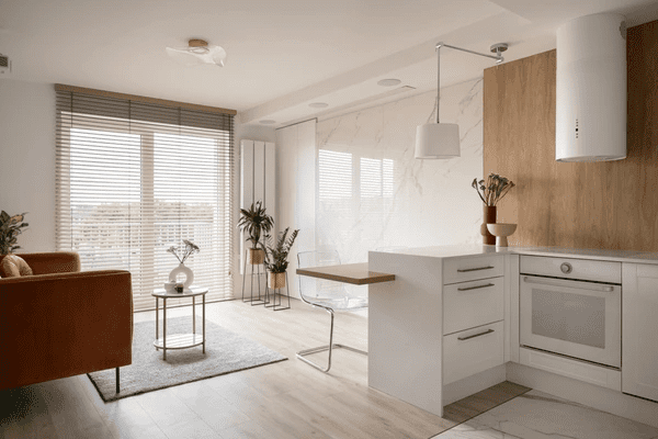 A stylish and modern apartment interior featuring sleek wooden blinds, a cozy brown sofa, a minimalist white kitchen, and natural decor elements.