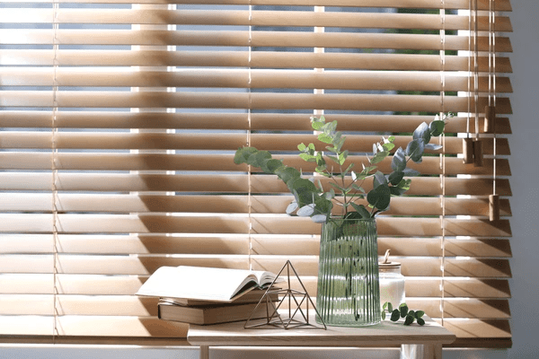 Elegant wooden window blinds filtering sunlight, with a decorative vase, open book, and geometric decor on a wooden table.