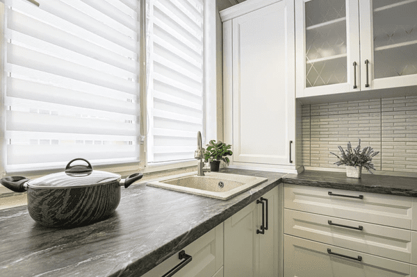 A modern kitchen featuring stylish Roman blinds 2025, offering privacy and elegance with a black marble countertop, white cabinets, and a clean aesthetic in Dubai.