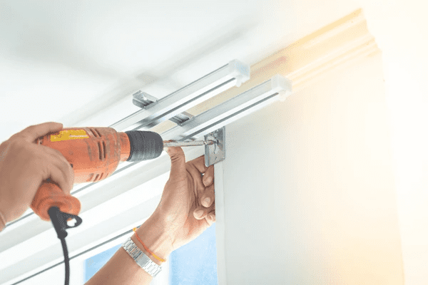 A close-up of a professional installer using a power drill to secure a curtain rail bracket above a window.