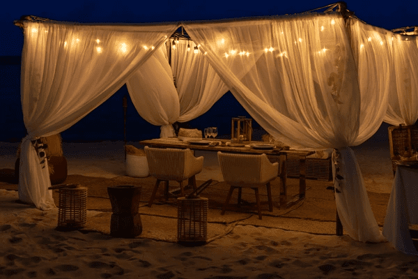 A stunning outdoor beachside dining setup with flowing white sheer curtains, warm string lights, and elegant décor, creating a romantic and luxurious ambiance for an unforgettable experience.