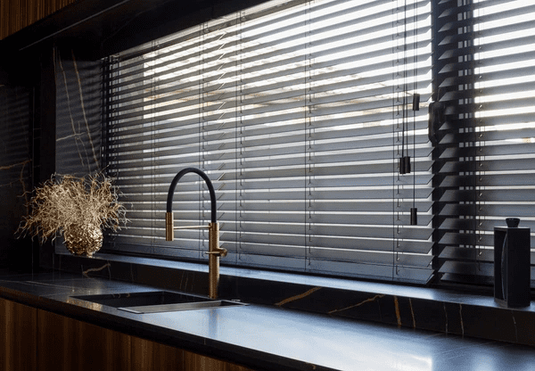 A stylish kitchen featuring black Venetian cut blinds, a black marble countertop with gold veining, a modern black and gold faucet, and minimalistic décor for a luxurious and sophisticated look.