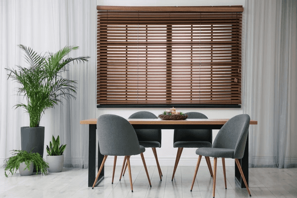 A contemporary dining room featuring a sleek wooden table, stylish gray chairs, lush green plants, and rich wooden cut blinds, creating a warm and modern ambiance.
