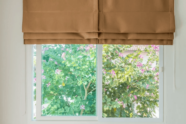 Elegant brown Roman blinds partially covering a large window with a view of lush greenery and blooming flowers, adding a trendy and stylish touch to the interior.