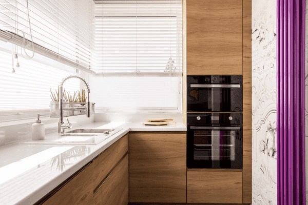 A contemporary kitchen with elegant wooden cabinets, a glossy white countertop, and modern white blinds covering the windows, offering a sleek and functional design—featuring the best kitchen blinds Dubai 2025.