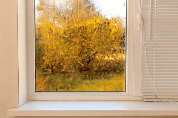 A modern window with moisture buildup and condensation, partially covered by white Venetian blinds, highlighting the need for proper ventilation and condensation-resistant blinds in Dubai 2025 homes.