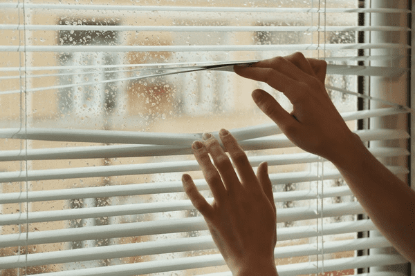 A person adjusting damaged white Venetian blinds on a rainy window, highlighting the impact of moisture buildup and condensation, and the need for durable, water-resistant blinds in Dubai 2025.