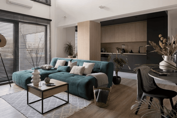 A contemporary open-concept living room with black soundproofing blinds, a plush teal sofa, and sleek wooden and black kitchen cabinets, creating a sophisticated and peaceful ambiance.