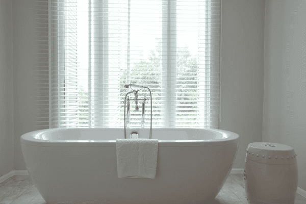 A minimalist bathroom featuring a freestanding bathtub with a sleek design, complemented by large windows covered with stylish white blinds for privacy.