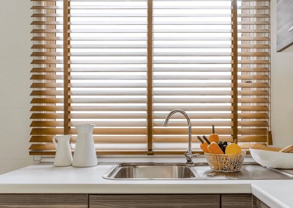 A modern kitchen with a sleek stainless-steel sink, a white countertop, and stylish wooden blinds filtering natural light. A fruit basket and white ceramic pitchers add a touch of warmth.