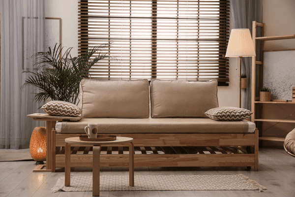 A stylish living room featuring a beige wooden sofa, a warm-lit floor lamp, and elegant wooden blinds covering a large window, creating a cozy and modern ambiance.