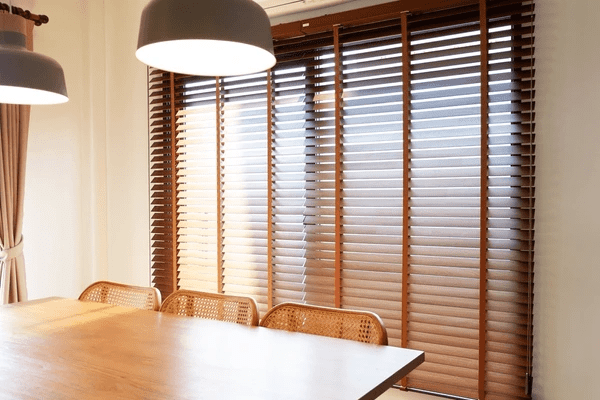 A modern dining room featuring a wooden table, woven chairs, and elegant wooden Venetian blinds that provide warmth, privacy, and energy efficiency.