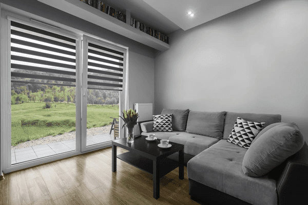 A modern living room with a stylish gray sectional sofa, a black coffee table, and sleek black-and-white zebra blinds on large glass doors overlooking a green landscape.