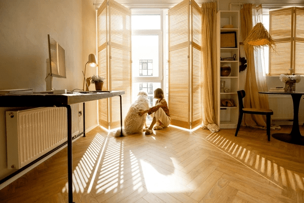 A cozy Dubai home featuring warm-toned wooden blinds, with a woman and her dog enjoying the soft natural light, showcasing pet-friendly blinds for modern interiors.
