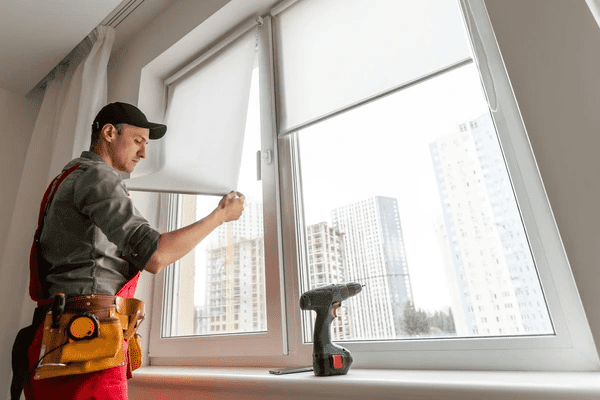 A skilled worker in a uniform installs white roller blinds on a modern apartment window, with a city skyline in the background.
