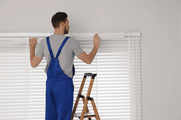 A professional worker in a blue uniform installing white horizontal blinds using a ladder, ensuring a precise and secure fit for a modern home in Dubai.