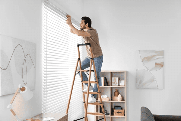A professional installer on a wooden stepladder adjusting white horizontal blinds in a modern, well-lit living room.