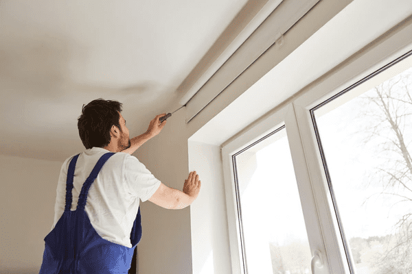 A professional installer in blue overalls fixing a roller blind above a large window in a bright modern room.