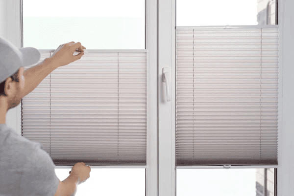 A man in a gray cap installing modern pleated blinds on a white-framed window for precise light control.