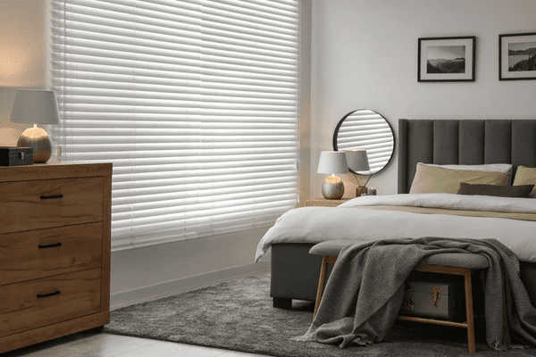 Cozy bedroom in Dubai with white horizontal blinds, grey upholstered bed, wooden furniture, and soft neutral tones.