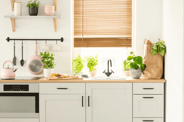 A modern white kitchen with wooden blinds, potted herbs, and natural sunlight—perfectly showcasing the best blinds for kitchens in Dubai 2025 with a cozy and stylish touch.