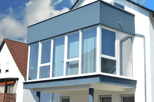 A contemporary upper-level conservatory with sleek vertical blinds inside large glass windows on a modern home exterior.