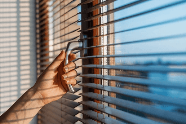 Close-up of a hand adjusting sleek wooden blinds on a sunny window, showcasing affordable and elegant blind options for Dubai homes in 2025.