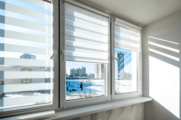 White zebra blinds installed on a sunny balcony window with a panoramic view of a snowy cityscape.