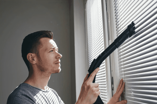 Man using a vacuum cleaner to clean dust off sleek fire resistant blinds in a stylish Dubai apartment.