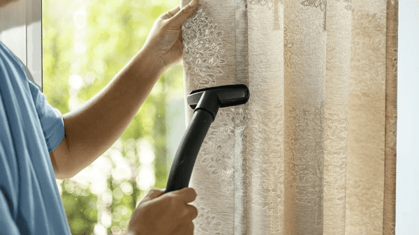 Close-up of a person vacuuming elegant patterned curtains near a bright window, highlighting cleaning and maintenance.