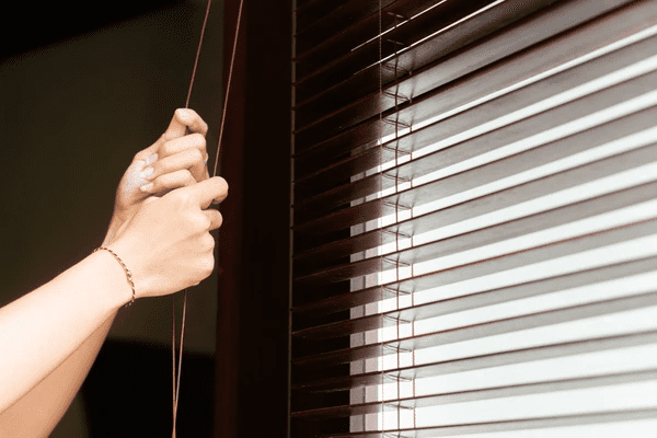 Hands adjusting brown wooden cord blinds, allowing natural light to enter through sleek, elegant window slats.