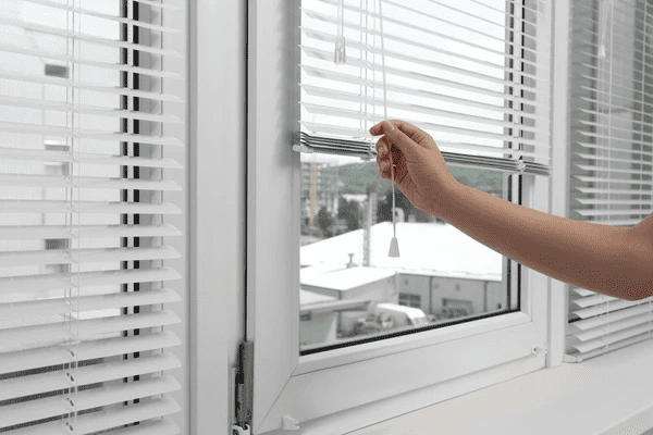 A person’s hand adjusting white horizontal cord blinds on a bright window inside a modern, cozy living space.