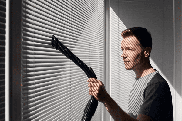 A man using a vacuum brush attachment to clean modern white blinds, ensuring a spotless and sleek look.