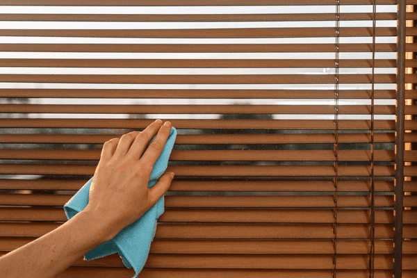 A close-up of a hand using a blue microfiber cloth to clean sleek brown wooden blinds.