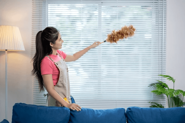 A woman in a pink shirt and beige apron dusting sleek white blinds in a bright modern living room.