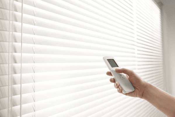 Hand operating motorized white blinds with a remote control in a bright modern living space.