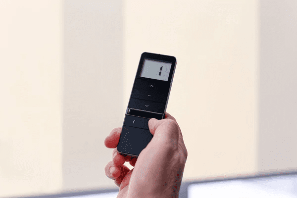Close-up of a hand operating smart blinds with a black remote control against a modern window backdrop.