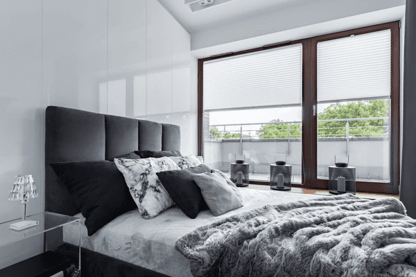 Modern bedroom with plush bed, cozy grey blankets, and privacy blinds on large balcony doors.
