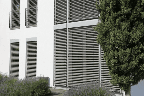 Contemporary white building with tall windows fitted with sleek grey horizontal outdoor blinds surrounded by greenery.