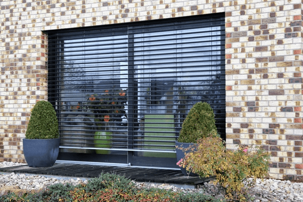 Black horizontal outdoor blinds on a large glass sliding door set in a brick wall, with potted greenery and modern landscaping.