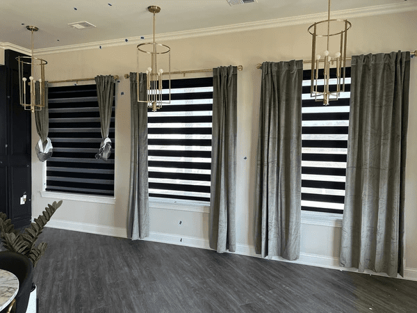 Elegant living room with black zebra blinds and grey velvet curtains on large windows, under gold pendant lights.